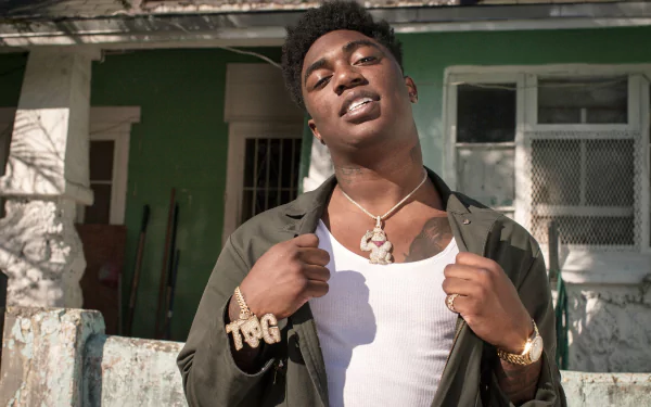 HD desktop wallpaper featuring a man in front of a house, wearing a necklace and adjusting his jacket.