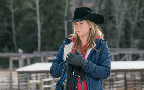A scene from the TV show Heartland featuring a young woman in a cowboy hat, holding ropes, set against a rustic farm background. This HD wallpaper captures the essence of ranch life.