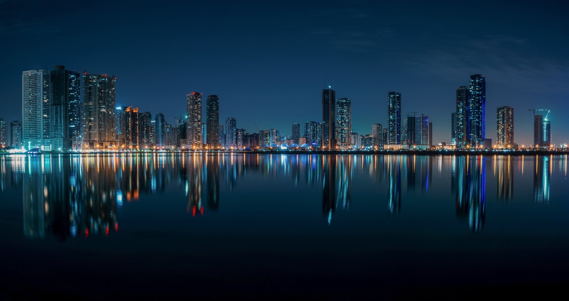 Night view of Sharjah, United Arab Emirates skyline with illuminated skyscrapers reflecting on calm water, captured in stunning 4K Ultra HD.