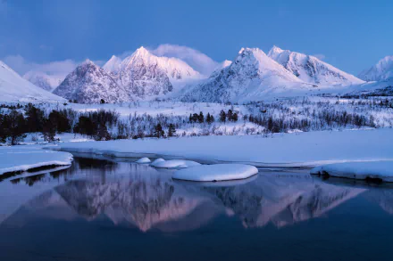 A tranquil winter scene in Norway featuring snow-covered mountains and their reflection in a serene lake. This HD wallpaper captures the pristine beauty of nature in winter.