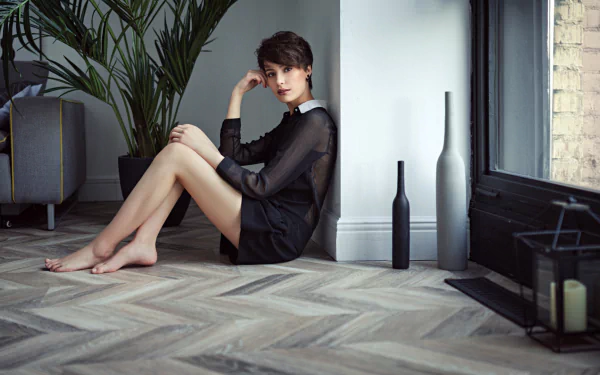 A woman with short hair in a black dress sits barefoot on a wooden floor near a window, surrounded by minimalistic decor, captured in a HD PC desktop wallpaper.