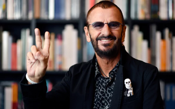 HD wallpaper featuring a smiling man flashing a peace sign, with a bookshelf background, representing Ringo Starr.