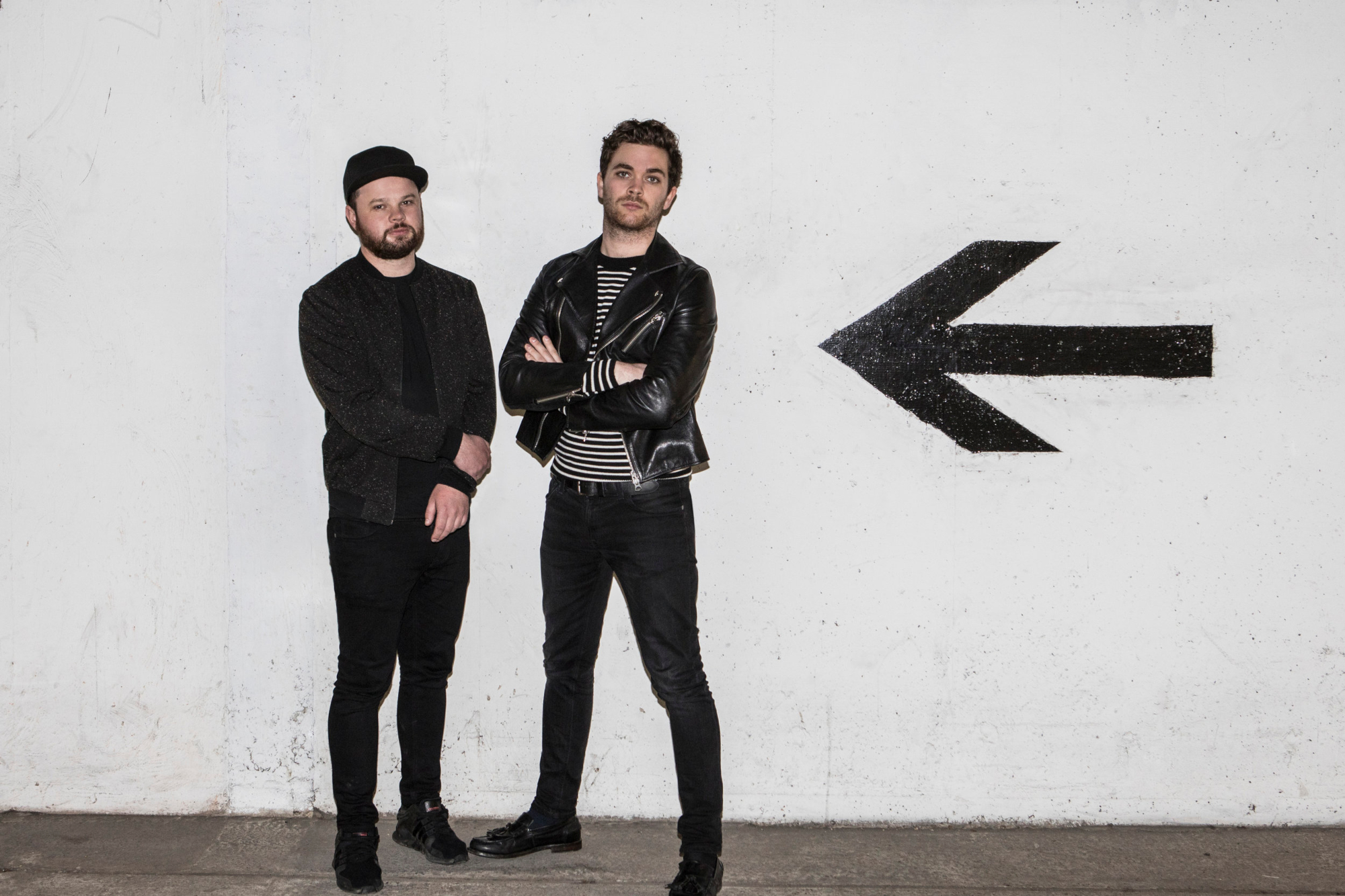 HD wallpaper of two band members from Royal Blood standing against a white wall with a large black arrow pointing left.