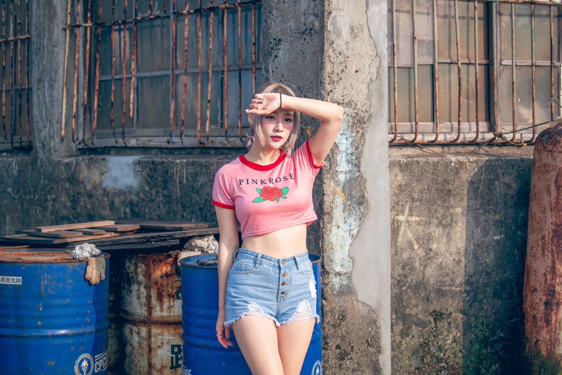 4K Ultra HD PC desktop wallpaper of a blonde model woman in a pink crop top and denim shorts standing by a weathered wall beside blue barrels.
