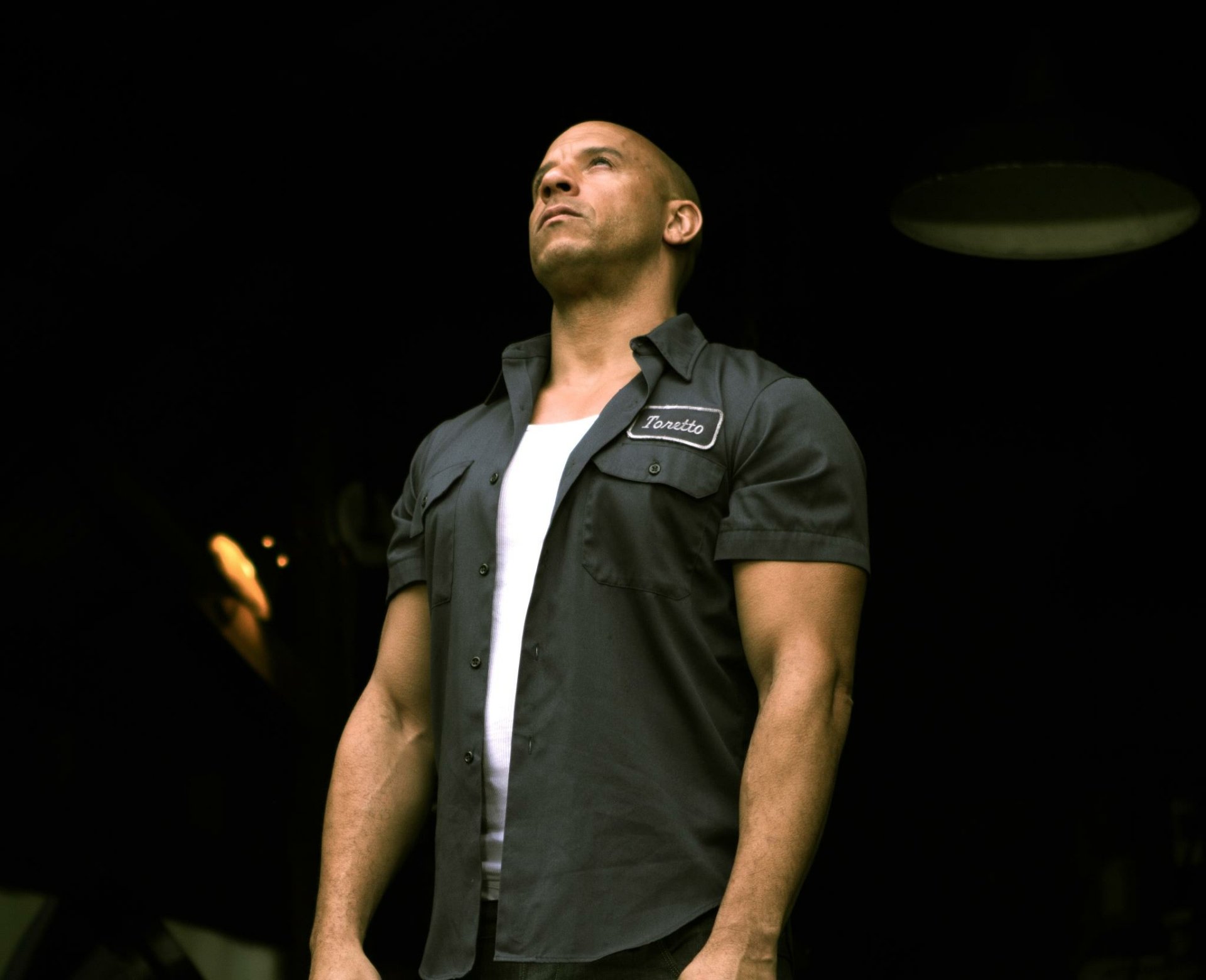 Vin Diesel stands confidently in a dark setting, wearing a black shirt over a white tank top, exuding a strong presence. This HD image serves as an eye-catching desktop wallpaper.