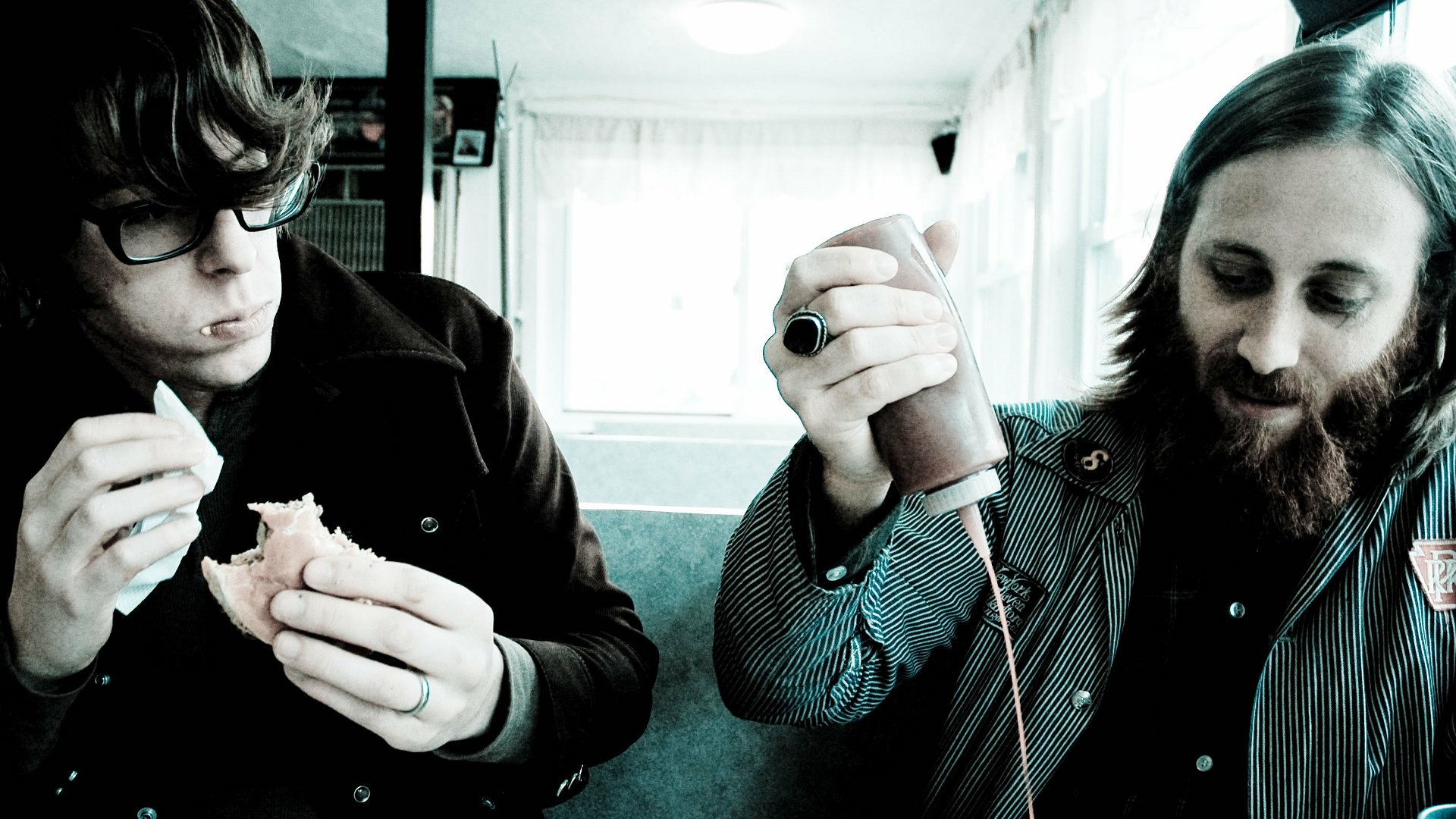 HD desktop wallpaper featuring two band members in a candid moment, one peering at a camera and the other engaged with a sandwich, reflecting the laid-back essence of rock duo.