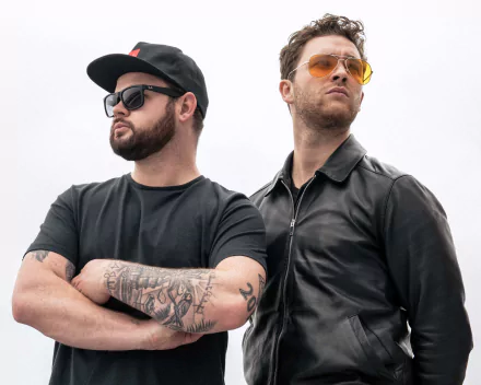 HD wallpaper featuring two confident men associated with Royal Blood, one with tattoos and a cap, the other with sunglasses and a leather jacket, against a clear background.