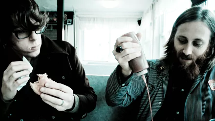 HD desktop wallpaper featuring two band members in a candid moment, one peering at a camera and the other engaged with a sandwich, reflecting the laid-back essence of rock duo.