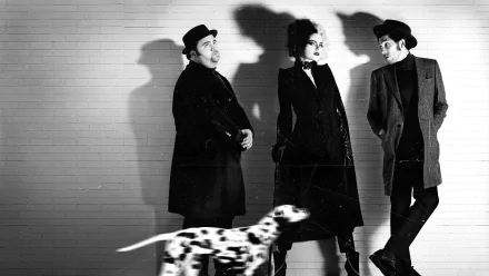 Black and white 4K Ultra HD wallpaper featuring Emma Stone as Cruella standing between two men against a brick wall with a Dalmatian walking in the foreground.