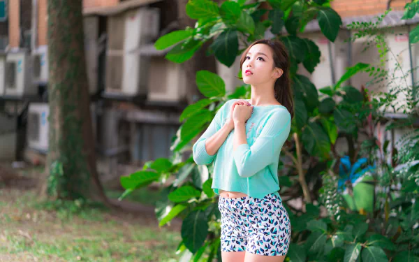 Asian brunette woman model wearing shorts and a light blue top stands outdoors near green foliage in a vibrant 4K Ultra HD desktop wallpaper.
