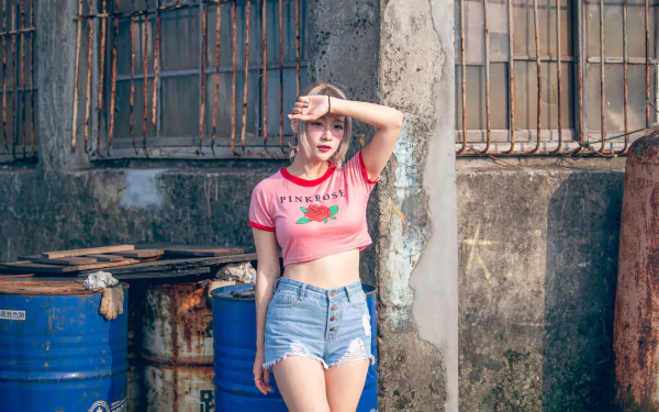 4K Ultra HD PC desktop wallpaper of a blonde model woman in a pink crop top and denim shorts standing by a weathered wall beside blue barrels.