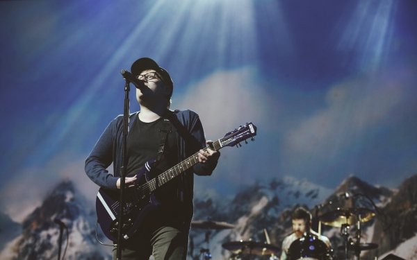 HD desktop wallpaper featuring a Fall Out Boy band member playing guitar on stage with dramatic lighting and a mountainous backdrop.