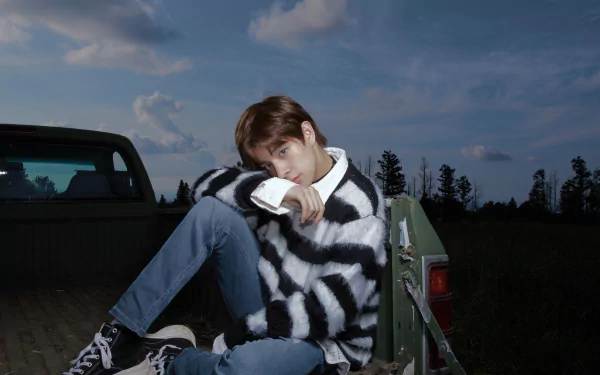 A captivating HD desktop wallpaper of a member from ENHYPEN, dressed in a striped sweater and sitting on a truck bed, against a backdrop of a twilight sky and serene landscape.