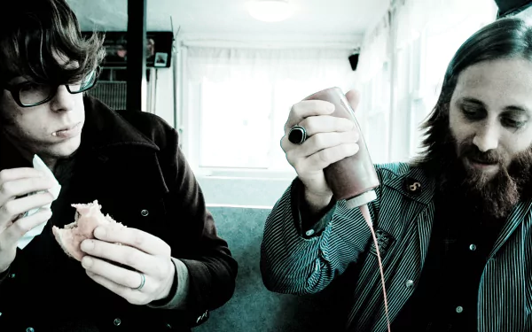 HD desktop wallpaper featuring two band members in a candid moment, one peering at a camera and the other engaged with a sandwich, reflecting the laid-back essence of rock duo.