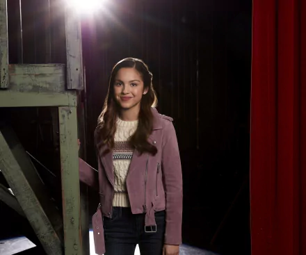 Olivia Rodrigo stands smiling on a stage set, featured in a HD desktop wallpaper from the TV show High School Musical: The Musical: The Series.