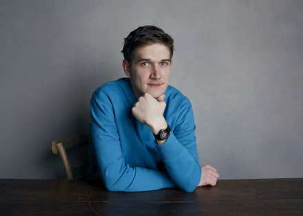 HD desktop wallpaper featuring a person in a blue shirt sitting at a table with a thoughtful pose, against a neutral gray background.