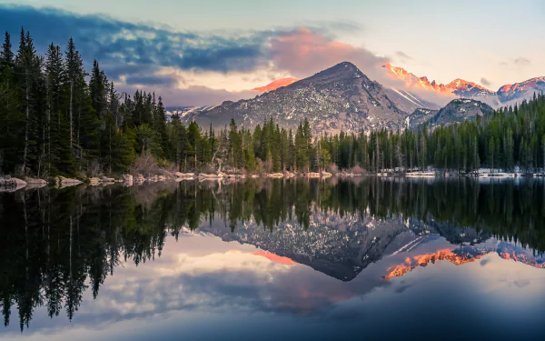 4K Ultra HD desktop wallpaper of the Rocky Mountains with a serene lake reflecting towering peaks and dense forest under a pastel sunset sky.
