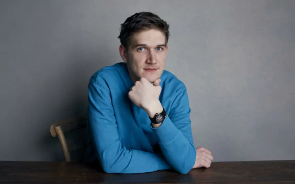 HD desktop wallpaper featuring a person in a blue shirt sitting at a table with a thoughtful pose, against a neutral gray background.