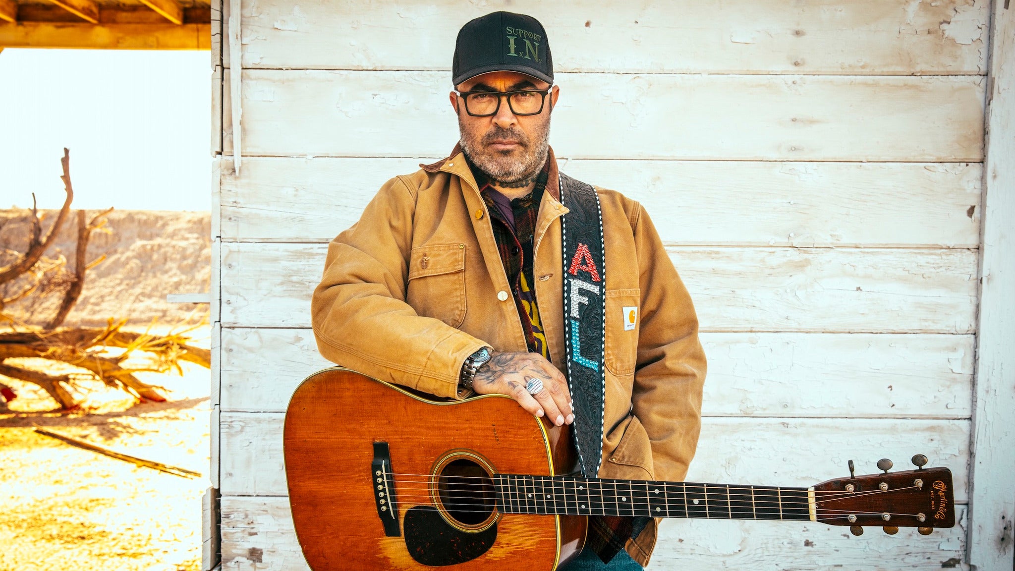 HD desktop wallpaper featuring a man with a guitar, wearing a brown jacket, cap, and glasses, posing against a rustic background.