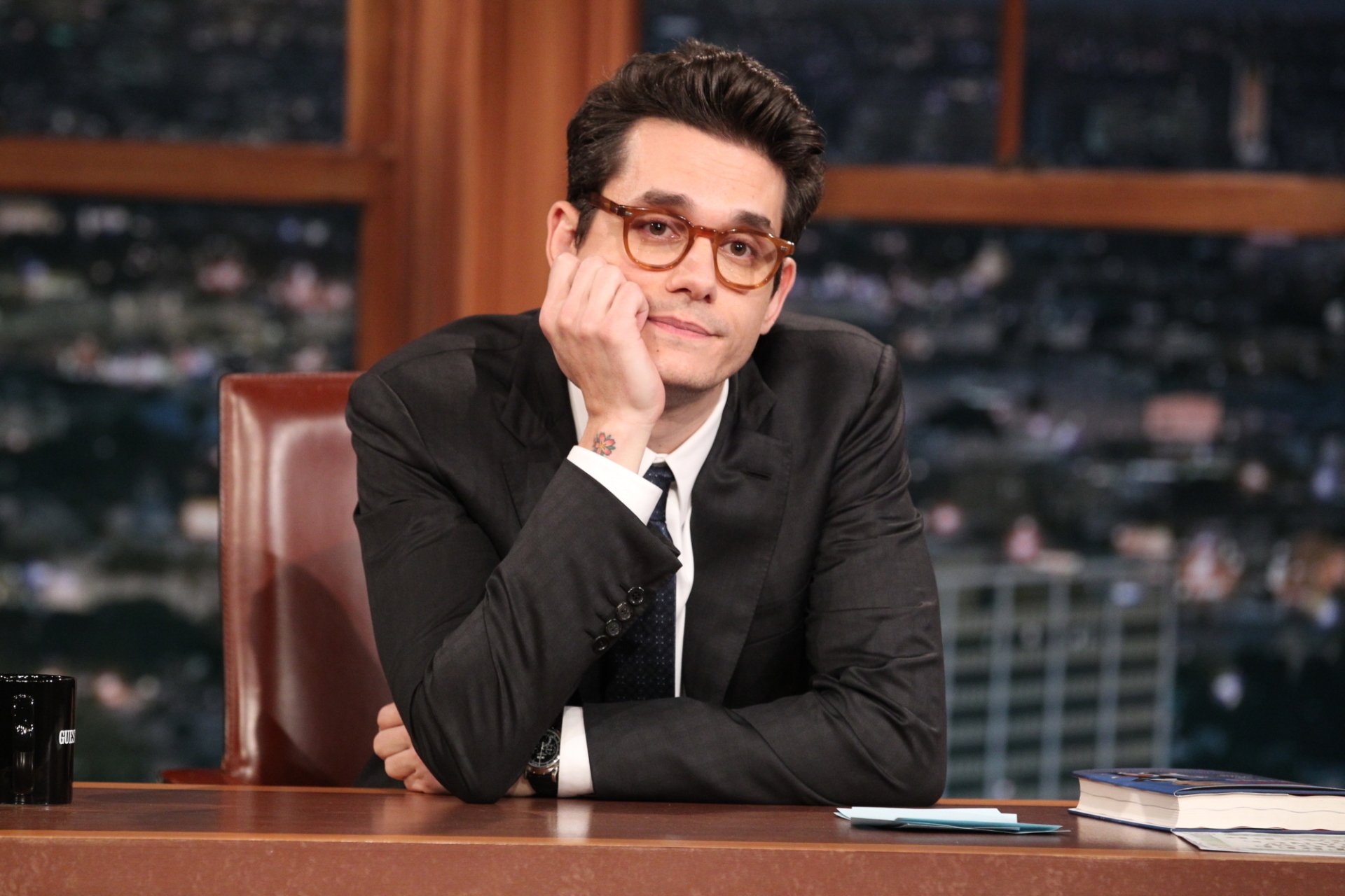 A high-definition desktop wallpaper featuring John Mayer seated at a desk, looking thoughtfully towards the camera, set against a nighttime city backdrop.