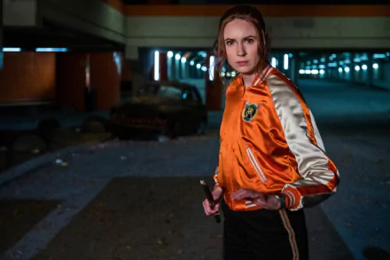 2K Quad HD PC wallpaper: cinematic film still from Gunpowder Milkshake — a determined woman in an orange satin jacket holding a weapon in a dimly lit parking garage.