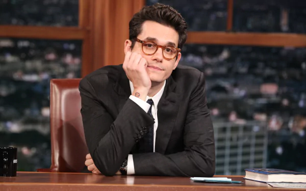 A high-definition desktop wallpaper featuring John Mayer seated at a desk, looking thoughtfully towards the camera, set against a nighttime city backdrop.