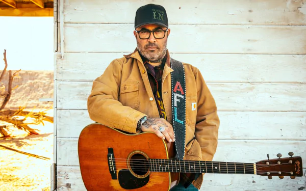 HD desktop wallpaper featuring a man with a guitar, wearing a brown jacket, cap, and glasses, posing against a rustic background.