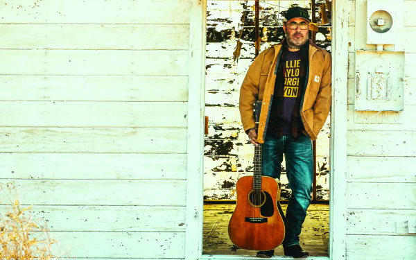 Man with guitar standing beside white wooden wall HD desktop wallpaper.