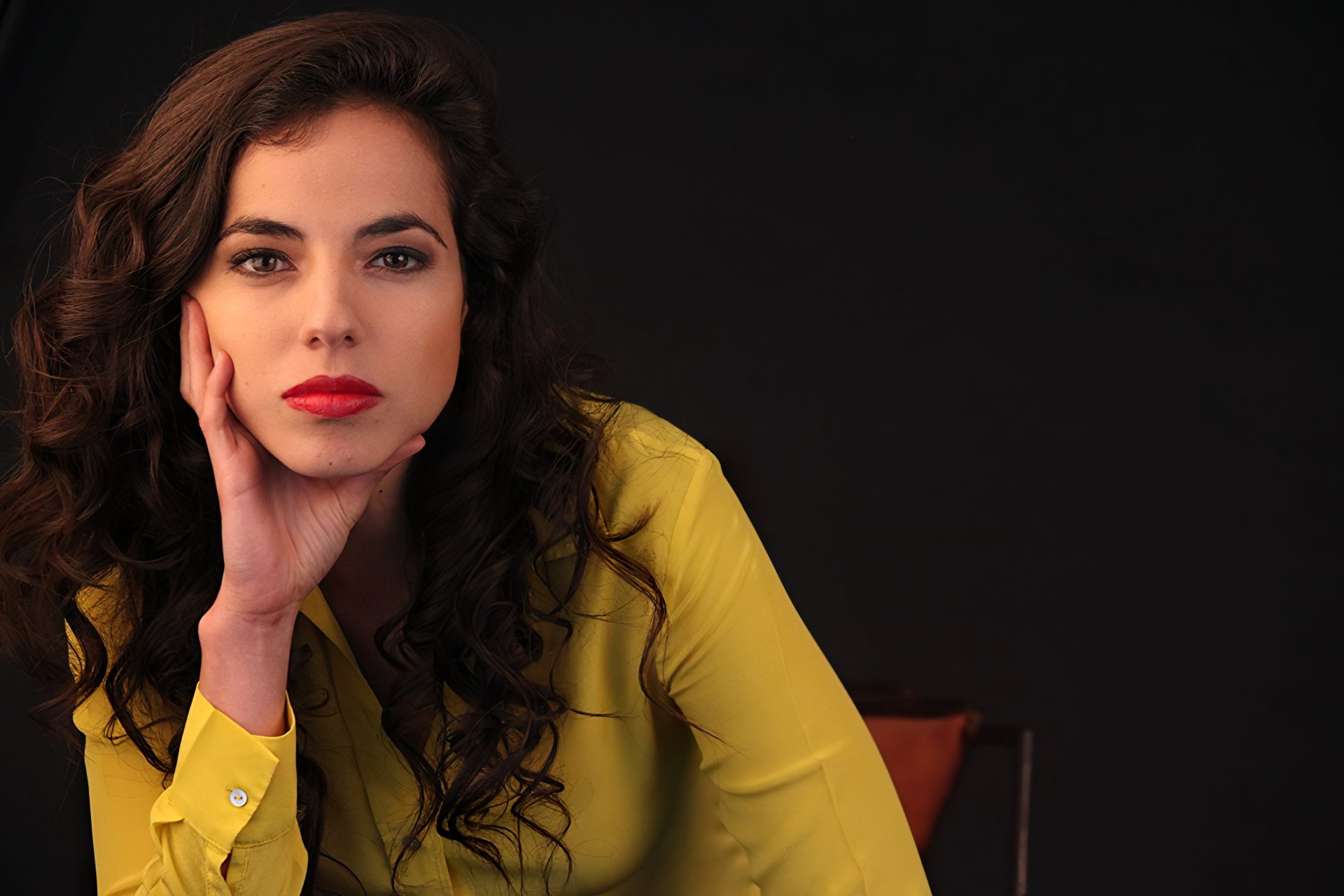 HD desktop wallpaper featuring a portrait of a woman in a yellow blouse leaning her cheek on her hand against a dark background.