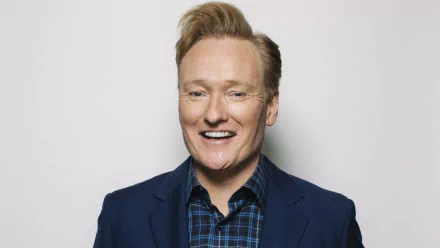 HD desktop wallpaper featuring a smiling man in a blue blazer and plaid shirt against a plain background.
