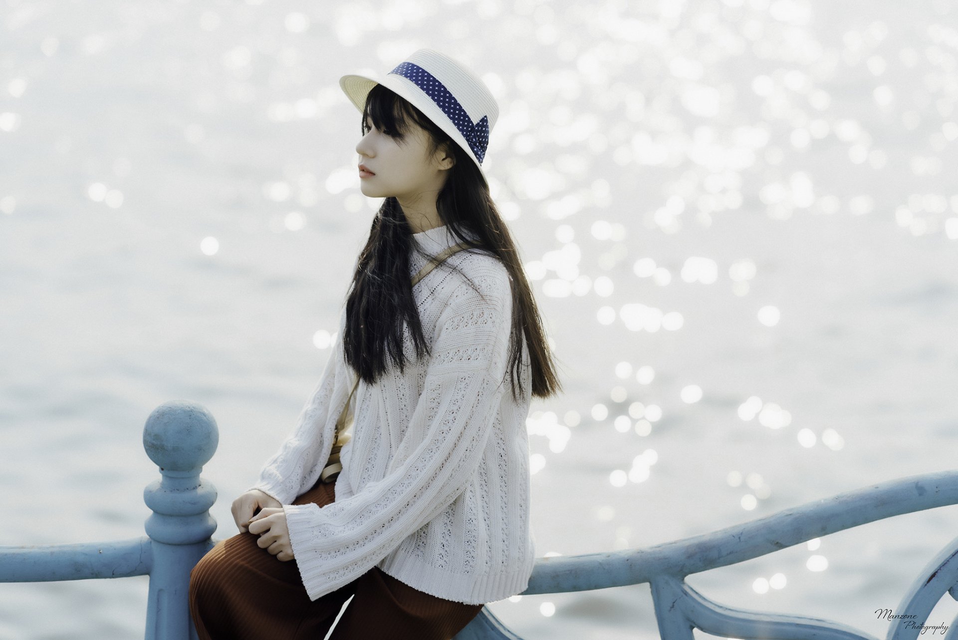 HD PC desktop wallpaper: a woman in a white sweater and straw hat sits on a pale blue railing, gazing over sparkling sunlit water.