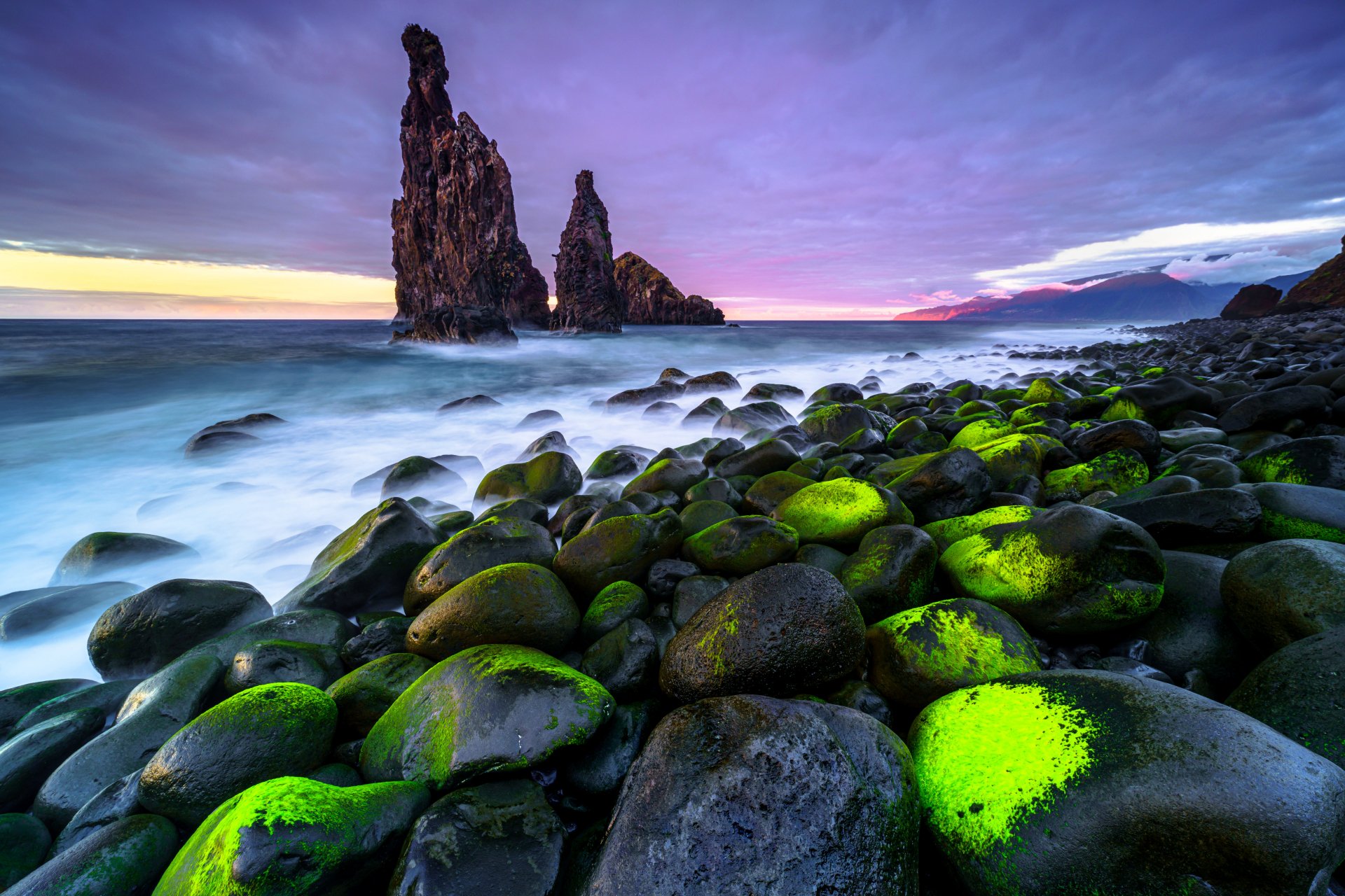 A stunning 4K Ultra HD seascape wallpaper featuring moss-covered stones along a rocky shore with towering sea stacks under a vibrant, cloudy sky at sunset.