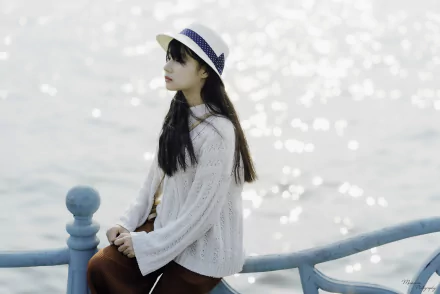 HD PC desktop wallpaper: a woman in a white sweater and straw hat sits on a pale blue railing, gazing over sparkling sunlit water.