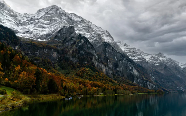 8K Ultra HD nature wallpaper featuring a serene lake reflecting autumn trees and towering snow-capped mountains under a cloudy sky.