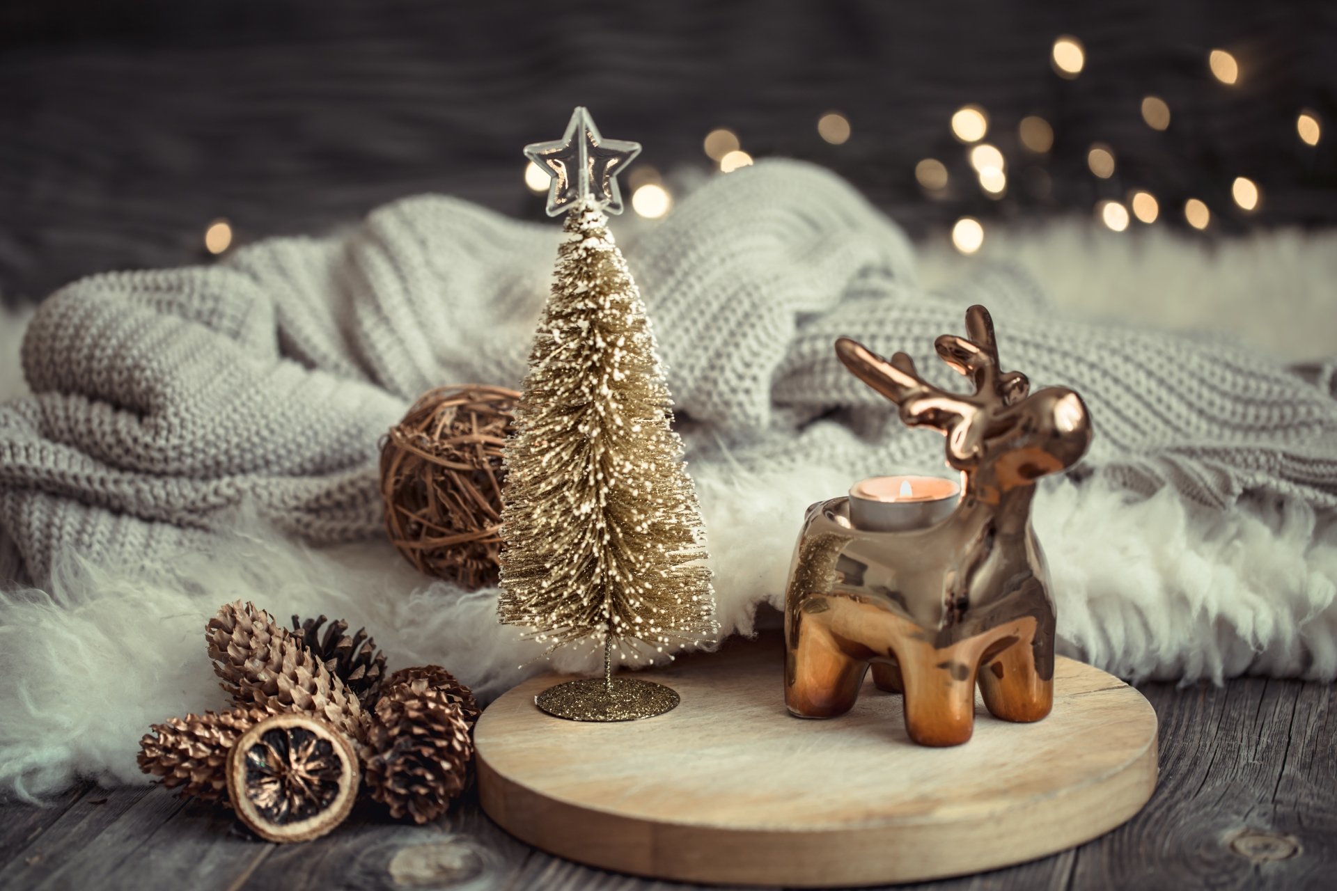 Cozy holiday scene of gold Christmas ornaments — a glittering miniature tree, bronze reindeer candle, pinecones and knit blanket with bokeh lights; 5K Ultra HD PC desktop wallpaper/background.