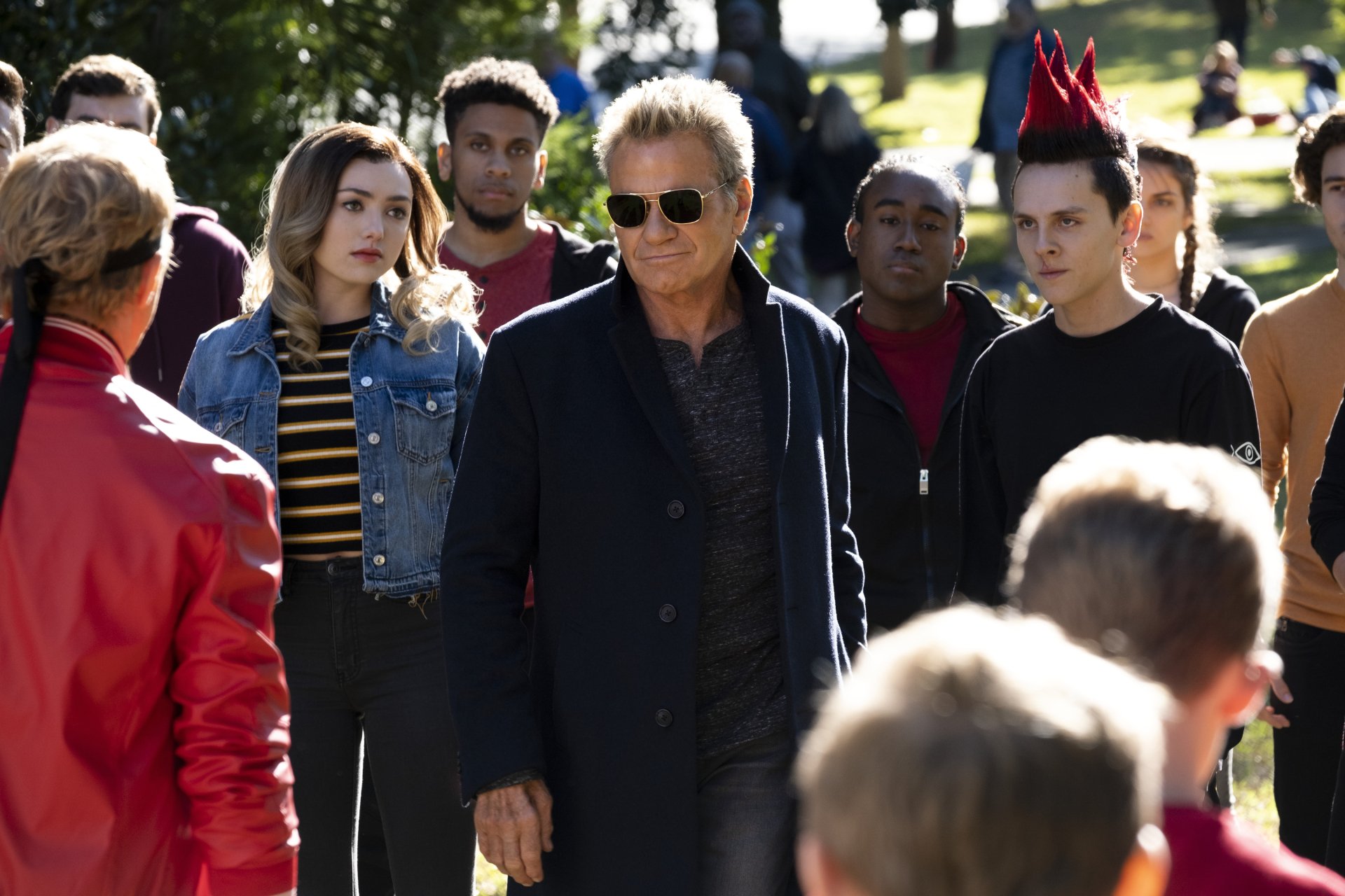 Cobra Kai TV show 2K Quad HD PC desktop wallpaper: a sunglasses-wearing man leads a tense outdoor standoff between rival students.