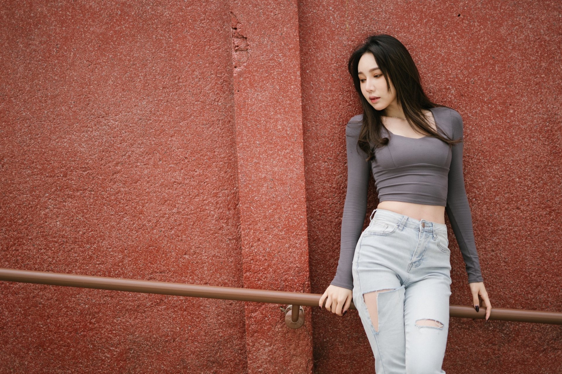 HD desktop wallpaper of an Asian woman with black hair wearing a gray long-sleeve top and ripped jeans, standing against a textured red wall with a railing.