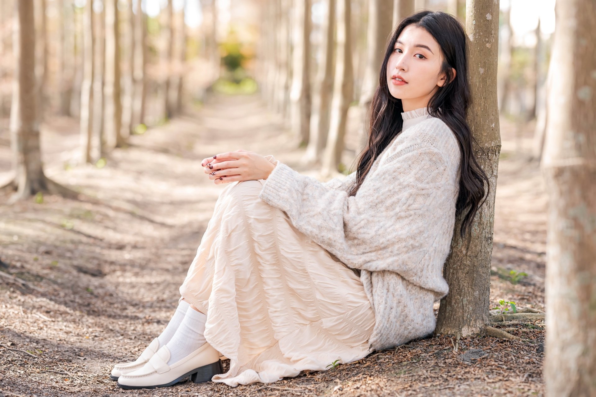 4K Ultra HD desktop wallpaper of an Asian woman with long black hair sitting against a tree in a sunlit forest, wearing a cozy sweater and flowing skirt.