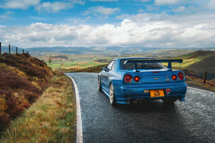A stunning HD wallpaper of a blue Nissan Skyline R34 parked on a winding road, surrounded by lush green hills and dramatic skies. Ideal backdrop for automotive enthusiasts.