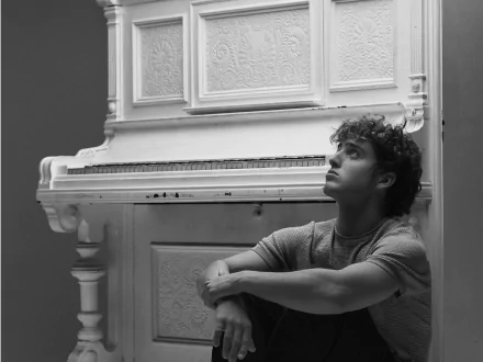 Black and white HD desktop wallpaper featuring musician Benson Boone sitting thoughtfully beside an ornate white piano.