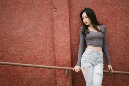 HD desktop wallpaper of an Asian woman with black hair wearing a gray long-sleeve top and ripped jeans, standing against a textured red wall with a railing.