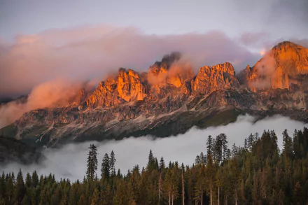 A stunning HD desktop wallpaper featuring the Dolomites with orange-tinted peaks at sunset, surrounded by clouds and lush forests below.