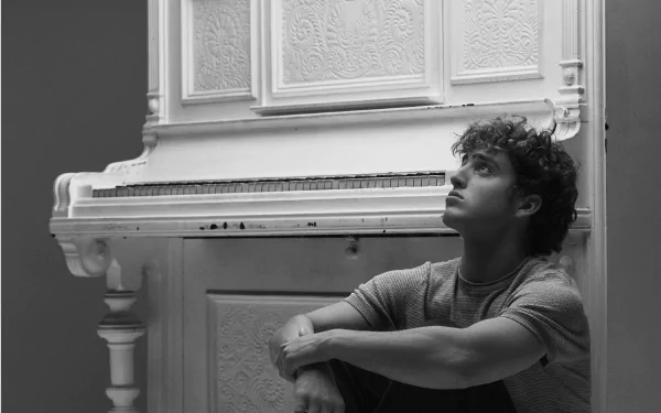 Black and white HD desktop wallpaper featuring musician Benson Boone sitting thoughtfully beside an ornate white piano.
