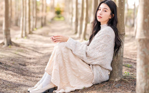4K Ultra HD desktop wallpaper of an Asian woman with long black hair sitting against a tree in a sunlit forest, wearing a cozy sweater and flowing skirt.