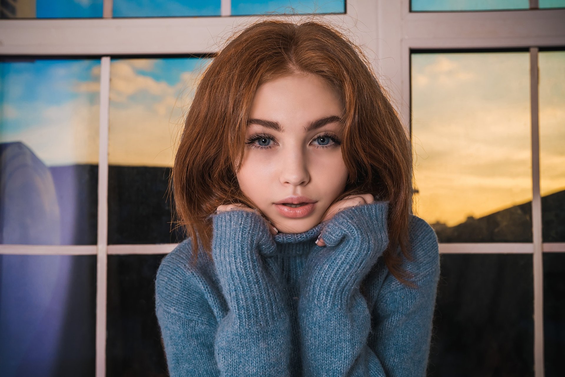 HD desktop wallpaper featuring a redhead woman model with striking blue eyes, wearing a cozy blue sweater, set against a window showing a sunset sky.