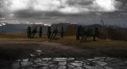 2K Quad HD PC desktop wallpaper: military artillery — a row of aged cannons on a barren field beneath brooding storm clouds, cracked stone foreground.