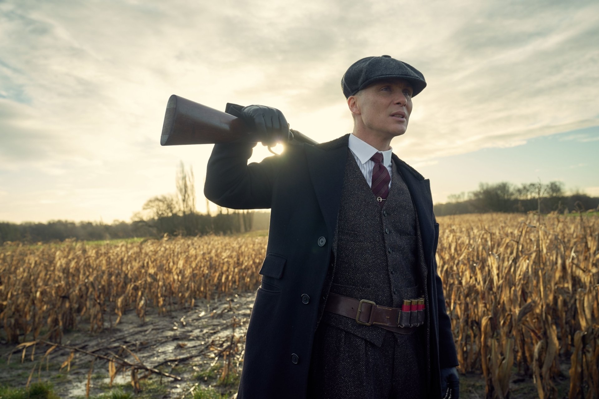 Peaky Blinders TV show 2K Quad HD PC desktop wallpaper: a man in a period suit and flat cap carries a shotgun over his shoulder while standing in a sunlit harvested field.