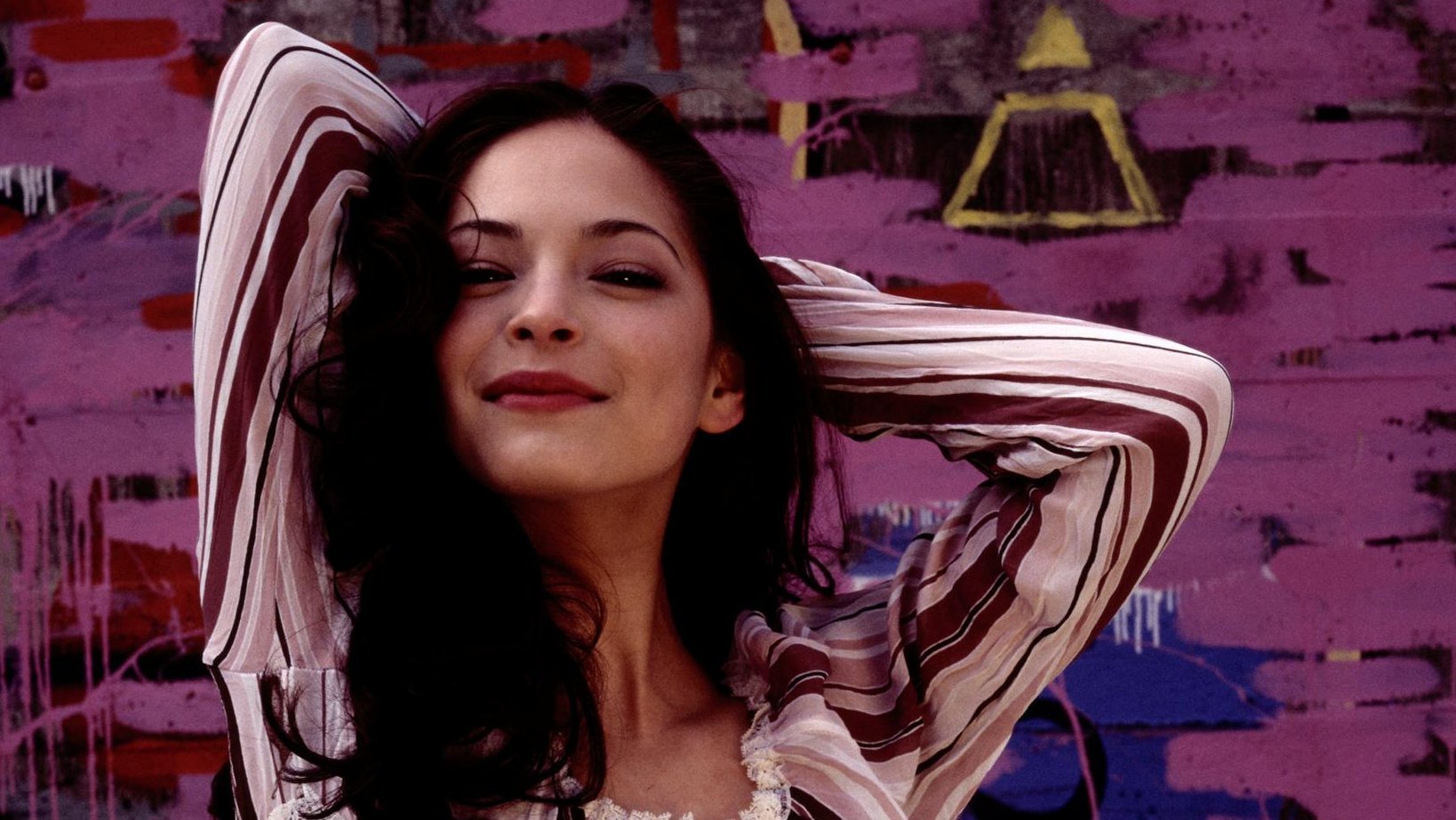 HD PC desktop wallpaper/background: smiling dark-haired woman in a striped top with hands behind her head, posed before a purple graffiti wall — identity not provided.