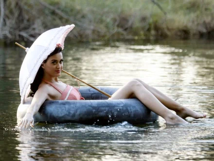 A singer with long black hair relaxes barefoot in a serene water setting, smiling while holding a pink umbrella, creating a vibrant and joyful scene.