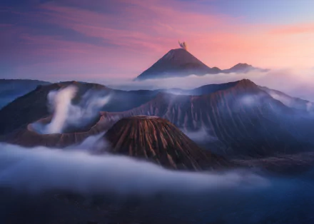 HD desktop wallpaper of Mount Bromo volcano rising above mist-filled calderas at sunrise, layered volcanic peaks beneath a pink-purple sky.